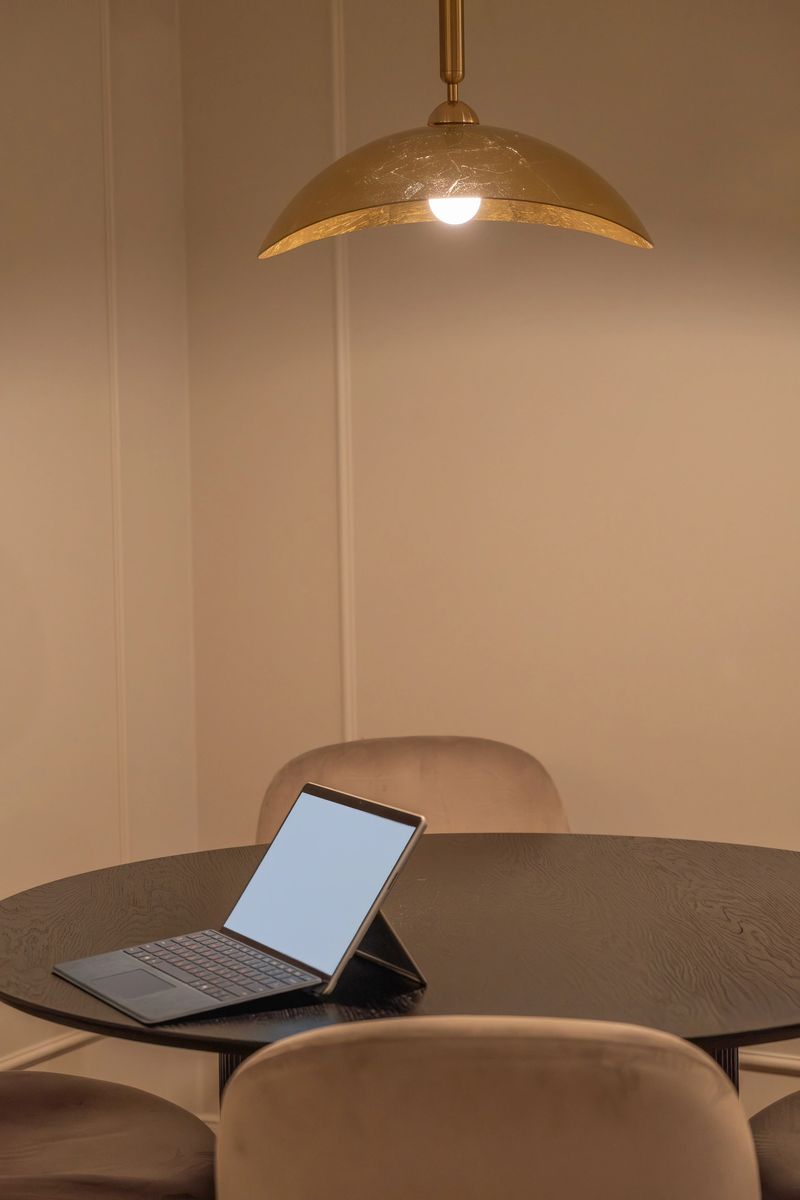 Modern workspace setup with an open laptop featuring a blank screen on a round dining table under a warm hanging lamp. Minimal interior with soft light and chairs creates a cozy home office atmosphere. Ideal for concepts of remote work, freelance lifestyle, online learning, business communication, digital technology, and copy space for mockup design.