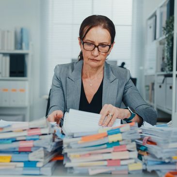 Woman in glasses sorting through a large stack of documents at a desk.