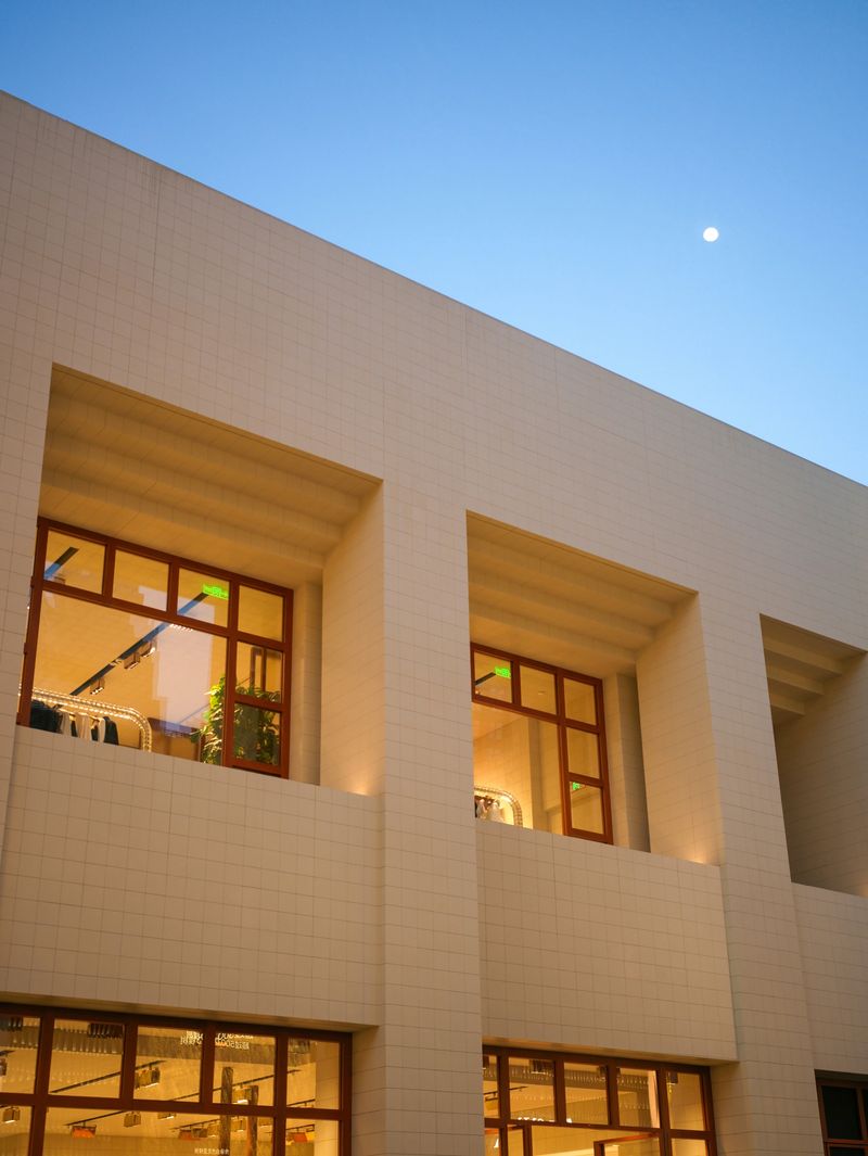 A minimalist modern white building facade featuring deep square cutouts that reveal warmly lit interiors, set against a calm twilight blue sky with a faint moon. Conveys contemporary residential or commercial design, warmth, and modern elegance.