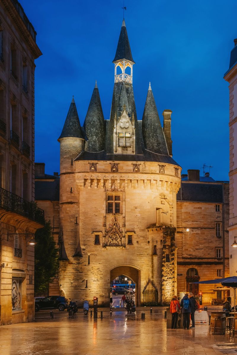 Illuminated medieval Porte Cailhau dominates a wet square at blue hour in Bordeaux, France, framed by historic buildings and a few pedestrians, evoking atmospheric nightlife, travel and architectural heritage.