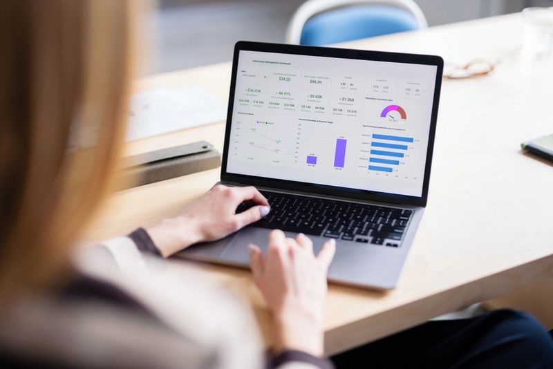 Businesswoman analyzes a financial dashboard on a laptop at a bright desk, focusing on charts and metrics.