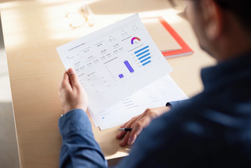 Business professional studies printed data report with charts and graphs at a bright office desk, concentrating.