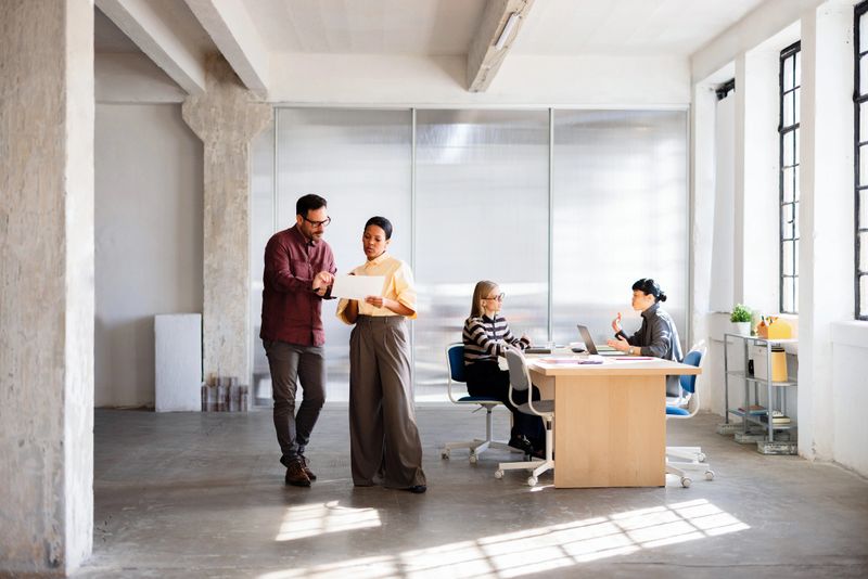 A diverse business team collaborates on documents and laptops in a bright, modern open-plan office space.