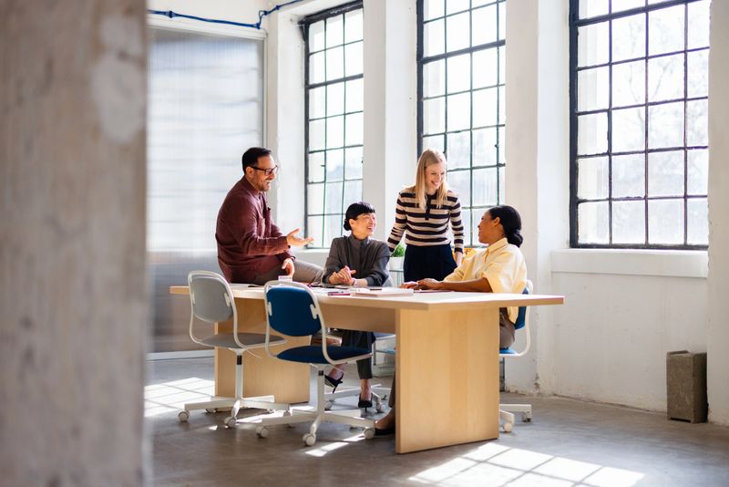 A diverse team brainstorming and smiling around a table in a sunlit, modern office space.