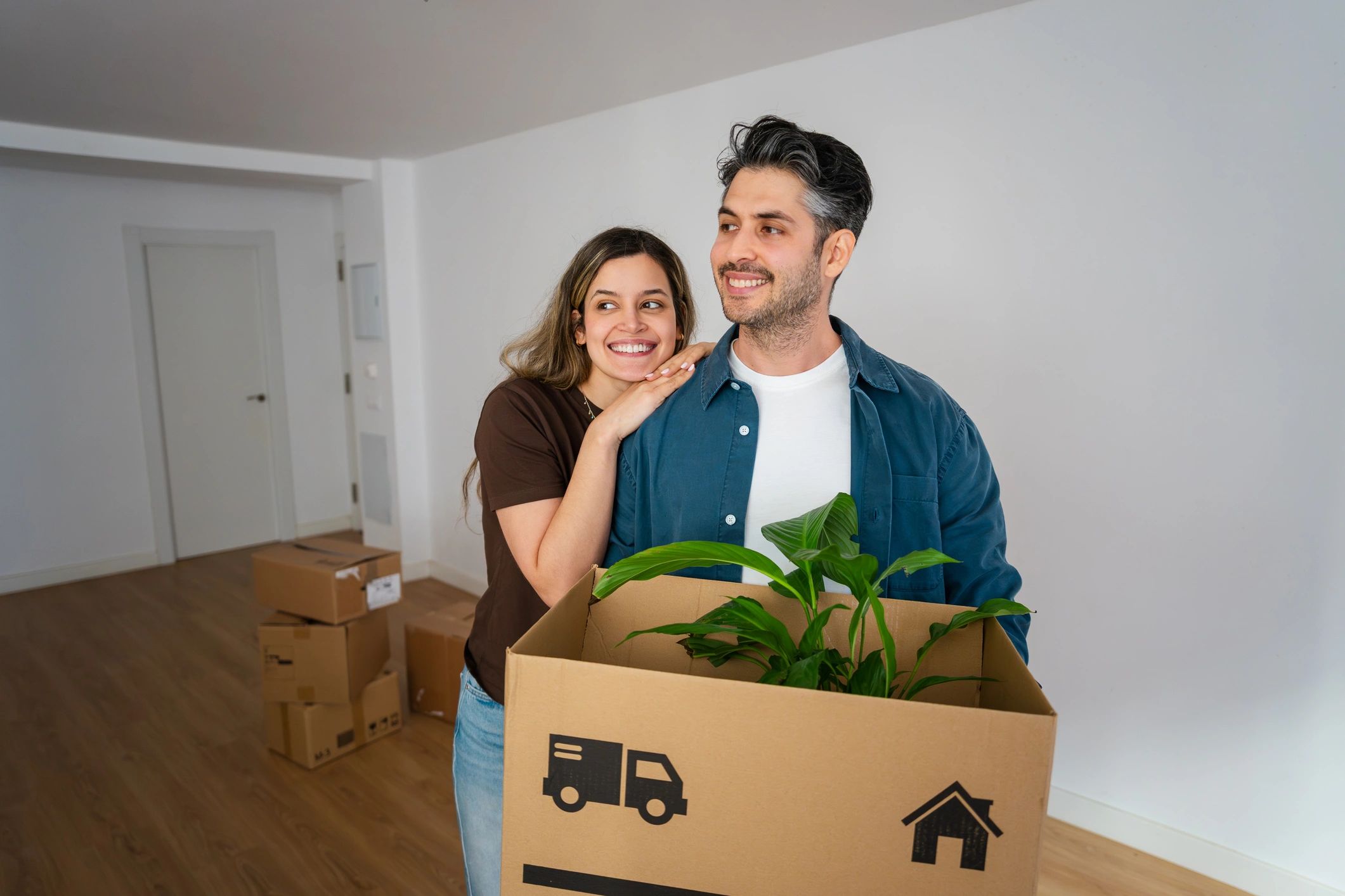 A smiling young couple stands inside their new, empty apartment during a move. The man holds a large