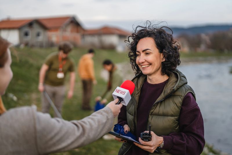 Smiling woman giving interview to reporter after a community river cleanup, volunteers collecting trash in background, promoting environmental stewardship and civic engagement