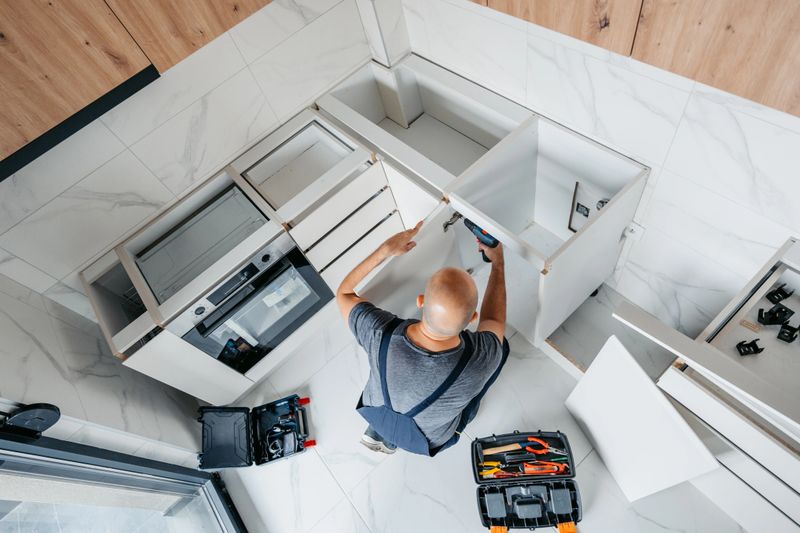 Mid-adult carpenter in overalls installing cabinets with a drill driver in an apartment.