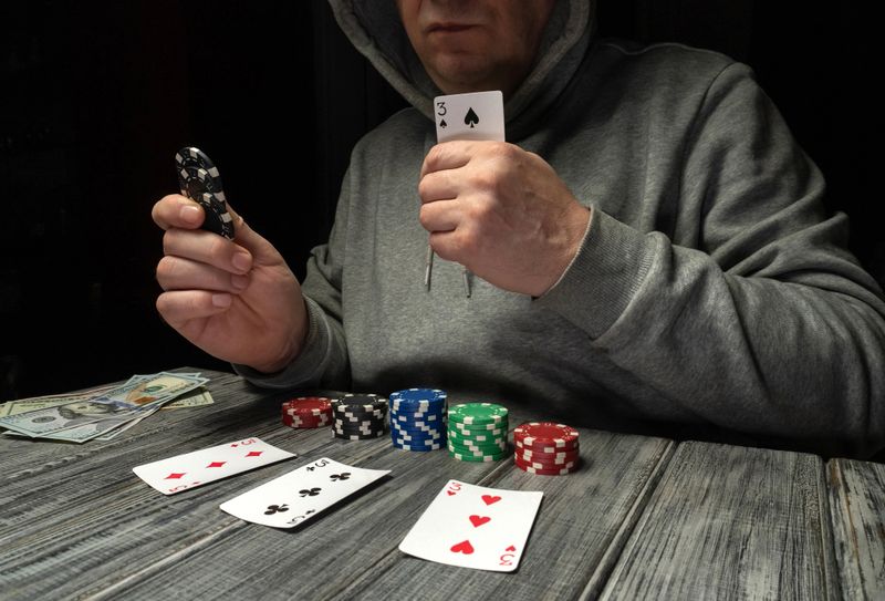 A man sits at a wooden table, holding a playing card in one hand and poker chips in the other. The cards are placed on the table while he concentrates on his next move in the game.