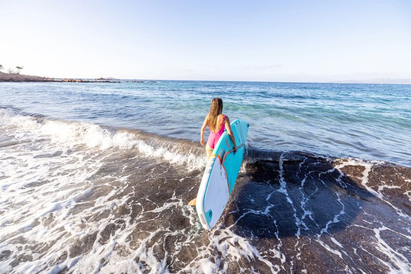 A woman walks into the sea carrying a paddle board, ready to begin her water adventure. The scene captures the spirit of summer, outdoor activity, and an active lifestyle by the ocean. Ideal for concepts such as paddleboarding, travel, freedom, and seaside recreation.