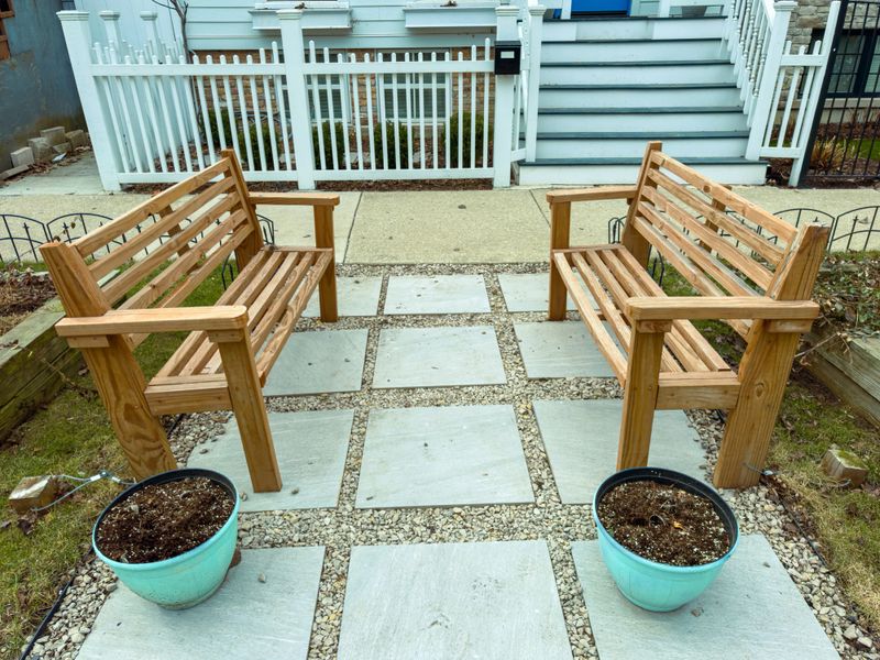 Two wooden benches are placed opposite each other on a path made of stones. There are planters near both benches. This setting is in a garden near a house.