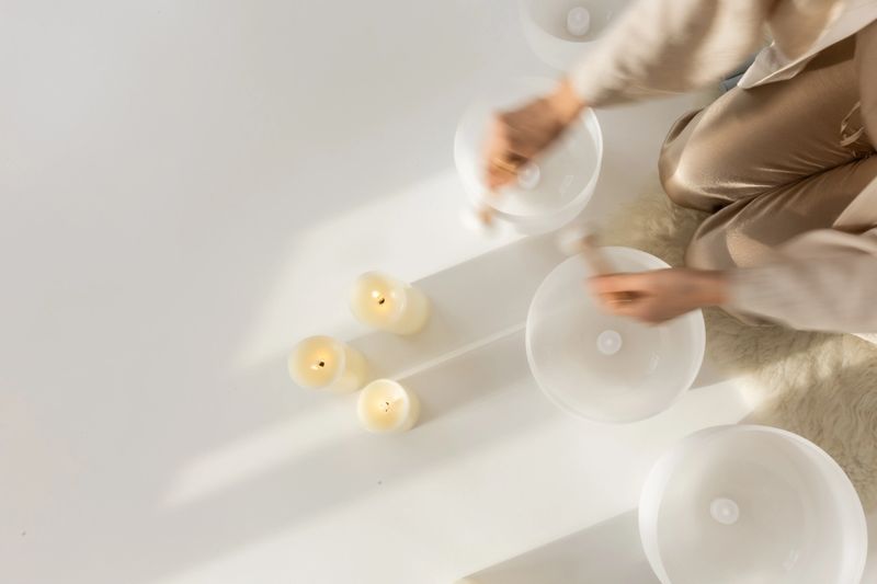 Overhead view of crystal singing bowls played in soft rhythmic motion, blurred hands and warm candles on white background. Concept of sound therapy, mindful relaxation, energy flow and holistic wellness.