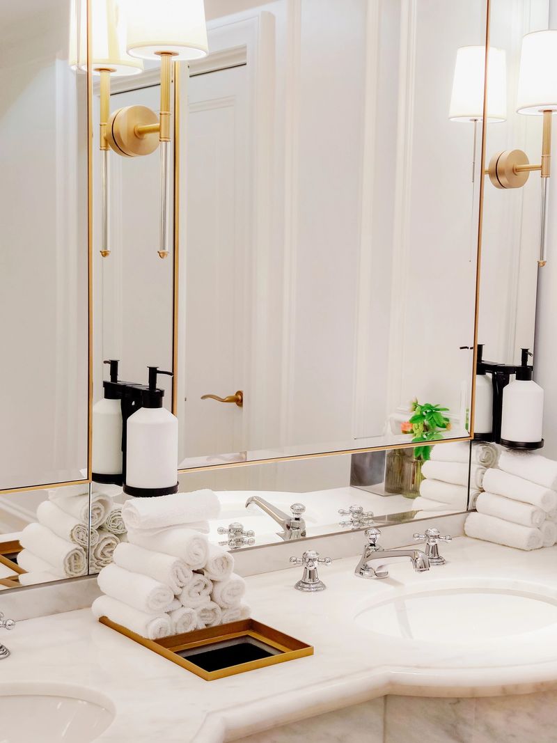 A beautiful, luxurious view of a neoclassical bathroom interior with wall sconces, large mirrors, a marble-topped vanity, white folded towels, and soap dispensers. Vertical close-up. Luxury lifestyle concept.