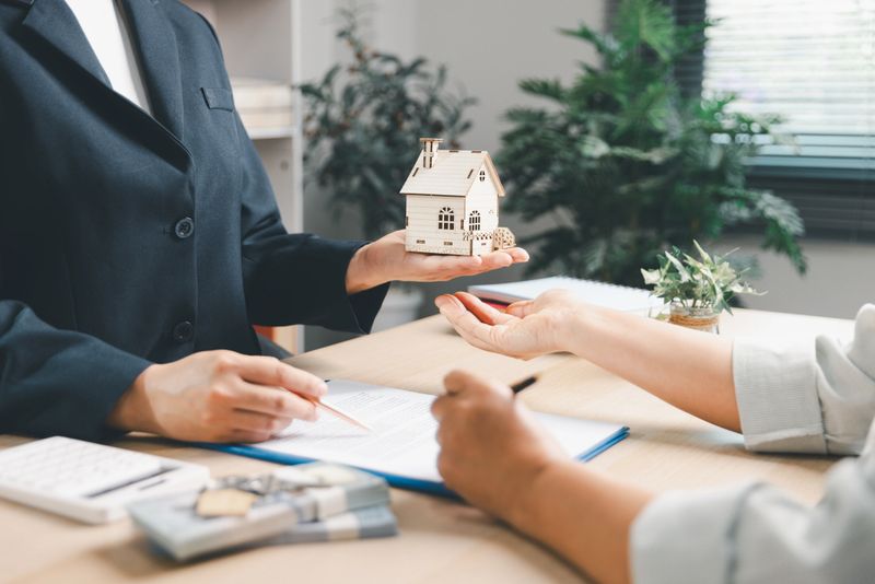 Professional real estate agent in a suit handing over a wooden house model to a client after signing a property contract. Desk features cash, keys, and documents for a successful home sale.