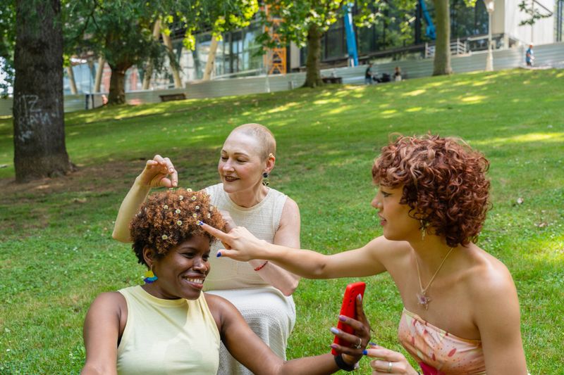 Three smiling diverse women friends decorating afro hair with small white flowers in a park on a sunny day
