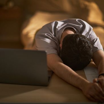Person exhausted, resting head on desk beside a laptop.