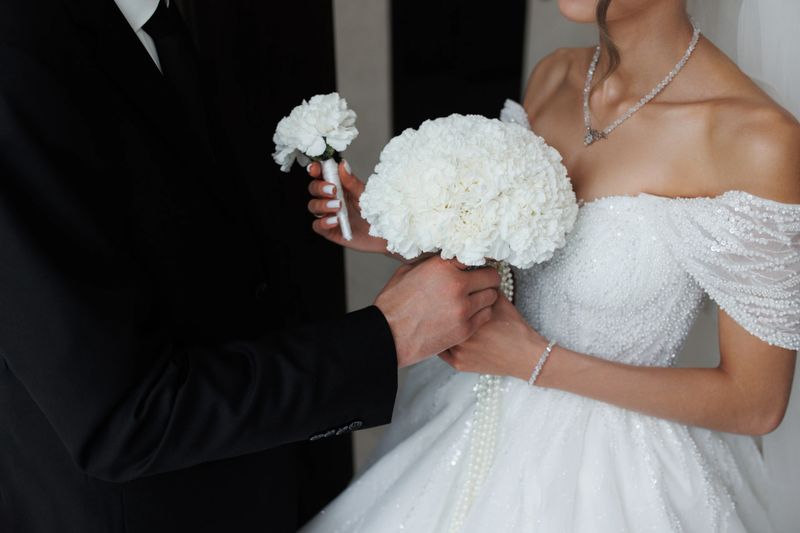 The bride and groom stand close, holding elegant white flower bouquets. The bride wears a shimmering gown while the groom is dressed in a classic black suit, capturing a romantic moment.