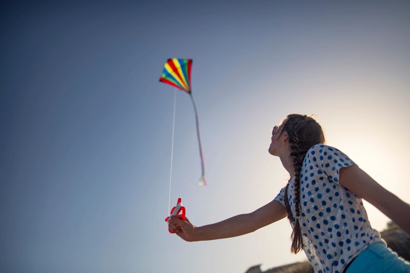 A low-angle shot looking up at a young girl as she expertly flies a rainbow-colored kite during a bright, sunny day. The girl, seen in profile with her hair in a long braid, holds the kite string handle firmly as she watches the kite soar high above. The composition emphasizes the expansive blue sky and the brilliant, backlit glow of the sun on the right, creating a feeling of freedom and optimism. This evocative image captures a quiet moment of focus and the simple, timeless joy of childhood outdoor play.Shot with Nikon D850