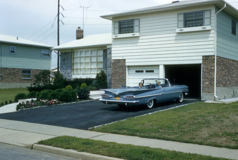 Vintage color photograph of a classic convertible parked in the driveway of a suburban mid century home with manicured lawn and garage, reflecting American suburban life in the 1960s.