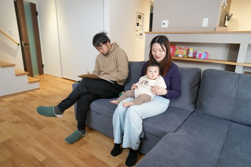 Cozy living room features a family moment. Mother holds baby while father writes notes. Both appear relaxed, focused on their shared activity. Soft lighting enhances warm, intimate atmosphere