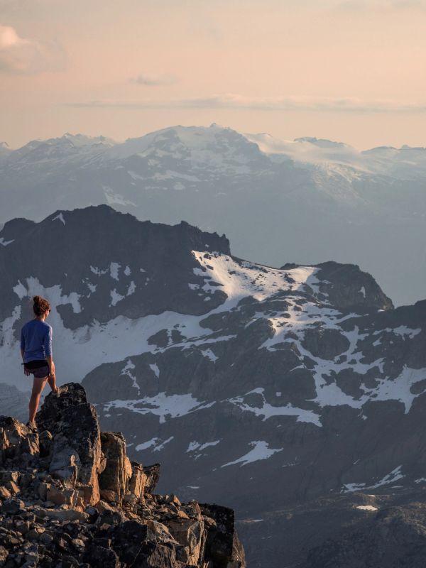 Person standing on rocky mountain peak with a snowy mountain range in the background.