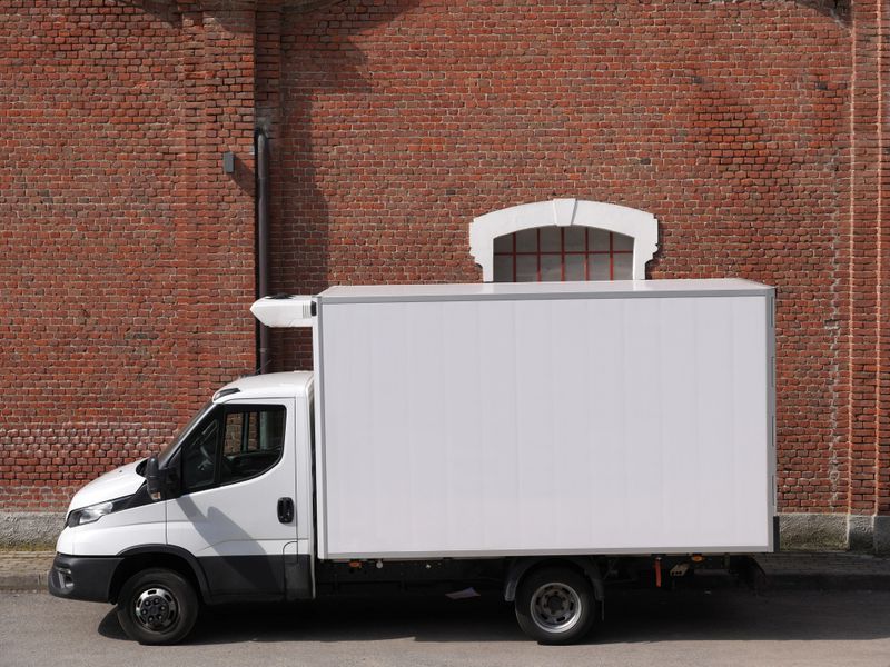 Side view of a white delivery truck parked against a brick wall, featuring a large blank panel ideal for advertising, branding or mockup use.