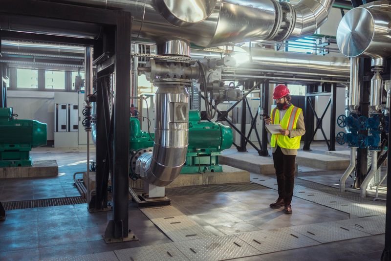 Male engineer wearing hard hat and safety vest checking machinery in an industrial power plant using a digital tablet