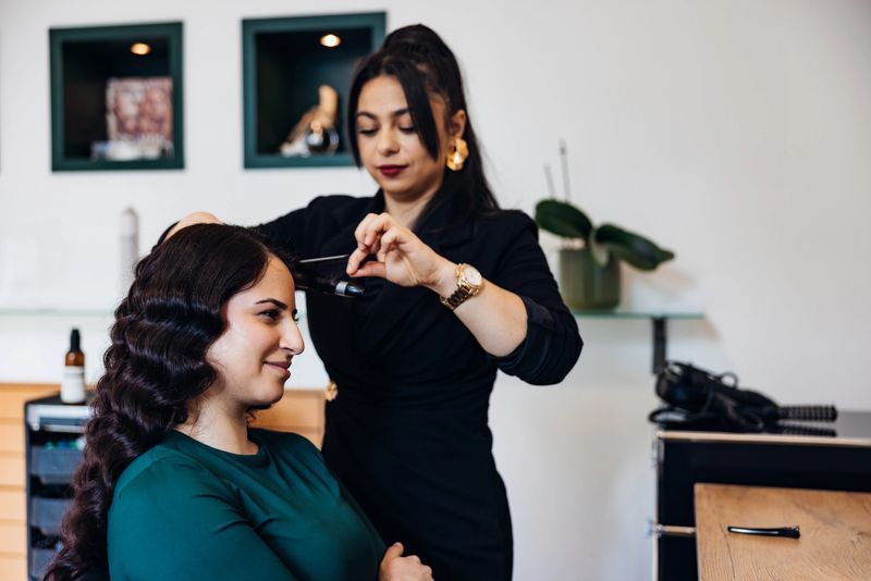 Professional hair stylist shaping a vintage finger-wave on a young woman's long dark hair in a modern salon, showcasing precision styling, elegance and client pampering