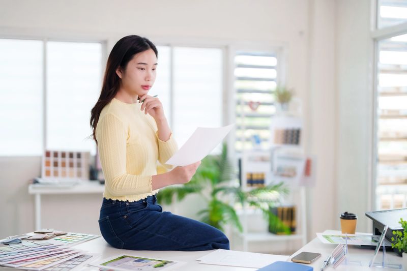 Asian adult woman professional evaluates design plans at bright modern workplace. Focused analyst contemplates important project blueprints.