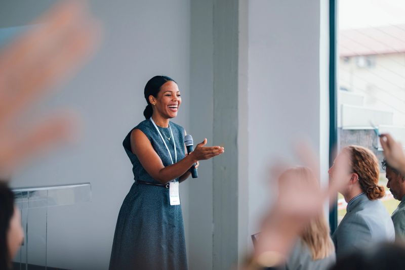 Confident female speaker presenting at a business conference workshop, engaging a seated audience with a microphone and name badge; a professional event focused on leadership, networking and education.