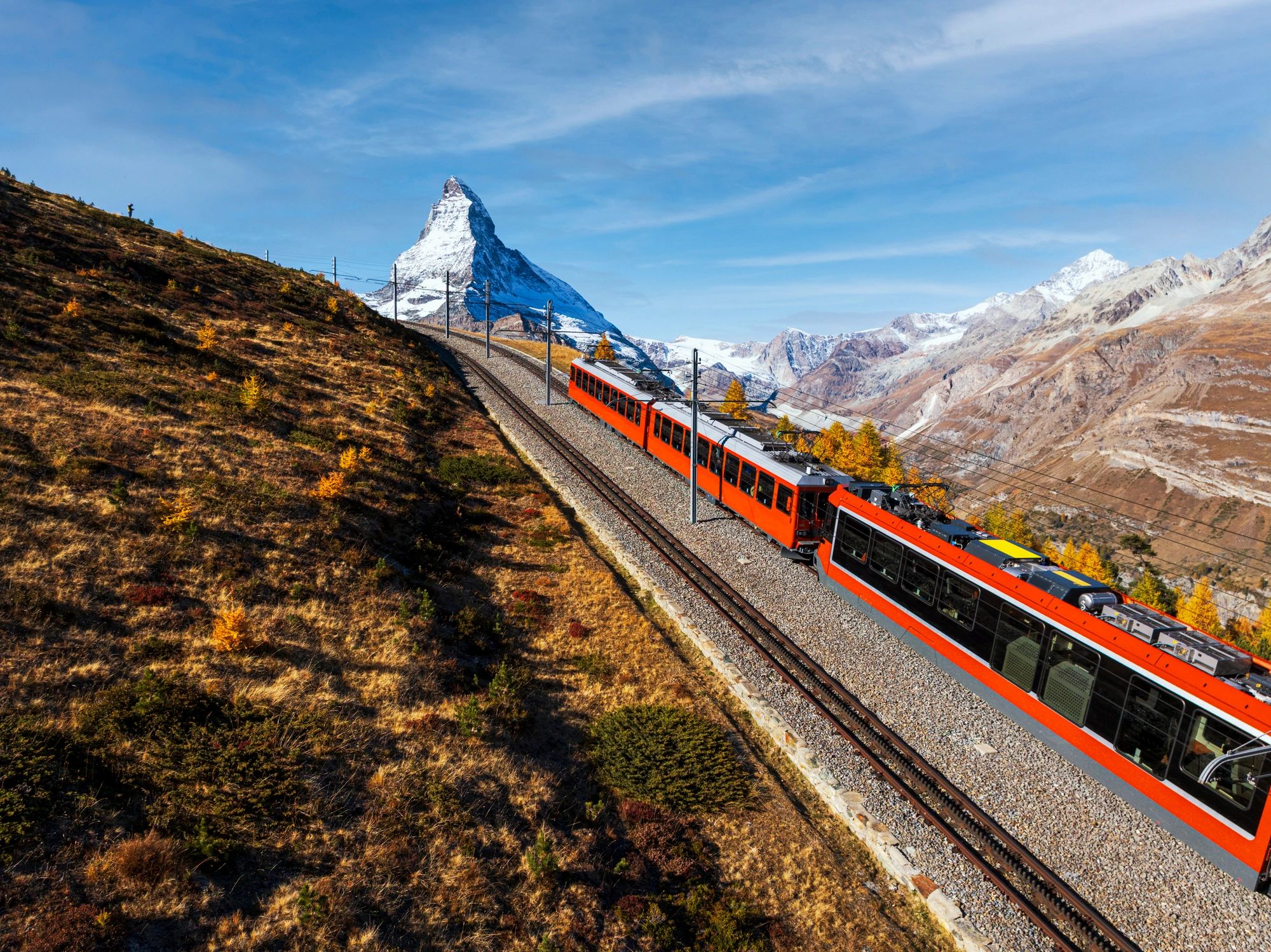 Red train climbs a mountain railway toward a snow‑covered mountain under a clear blue sky.