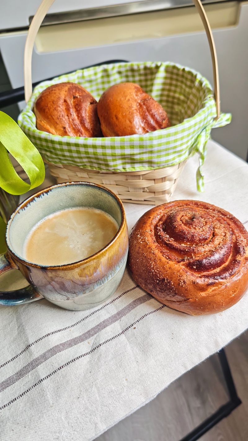 Freshly baked cinnamon rolls in a green checkered basket beside a cup of coffee on a linen tablecloth in a bright kitchen setting
