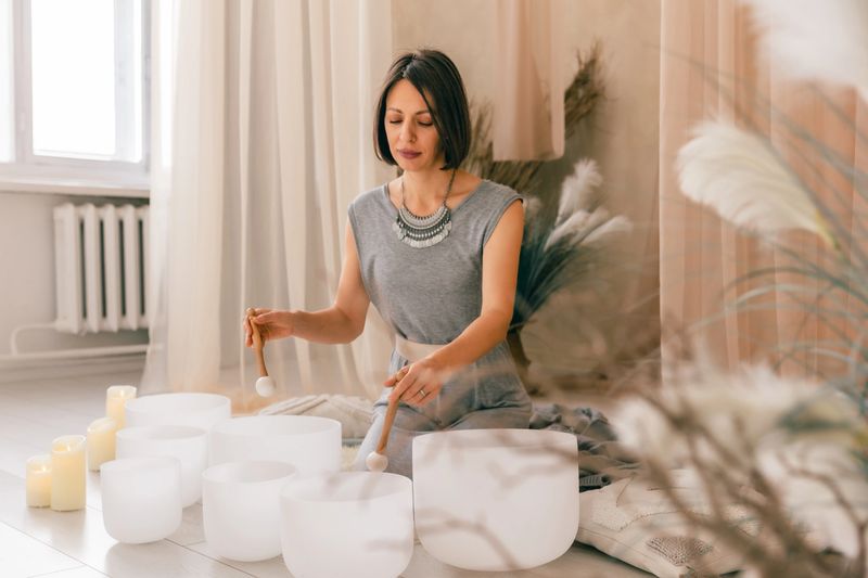 Mindful sound healing practice with a woman playing crystal singing bowls indoors. Calm atmosphere, soft candles, therapeutic vibrations, meditation, relaxation, and holistic wellness concept.