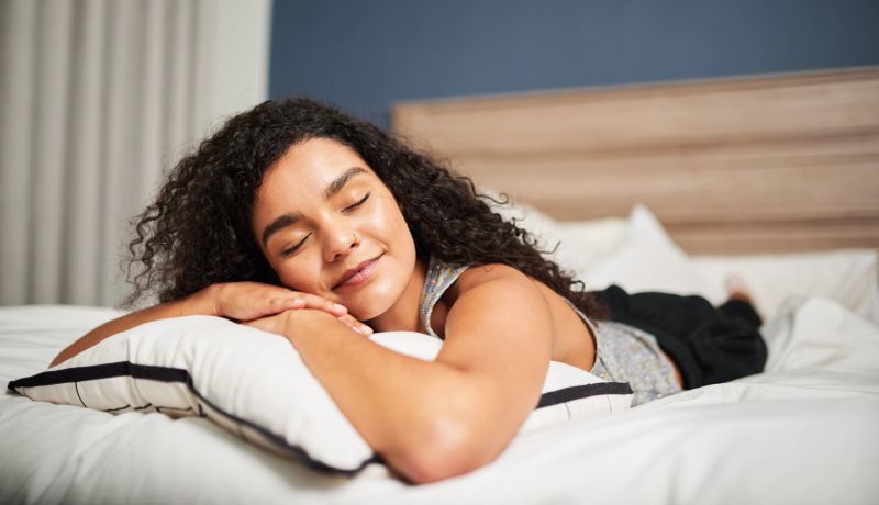 A relaxed young woman lies on her stomach on a bed with eyes closed, resting on a pillow in a bright, cozy bedroom conveying comfort, calm, and peaceful relaxation.