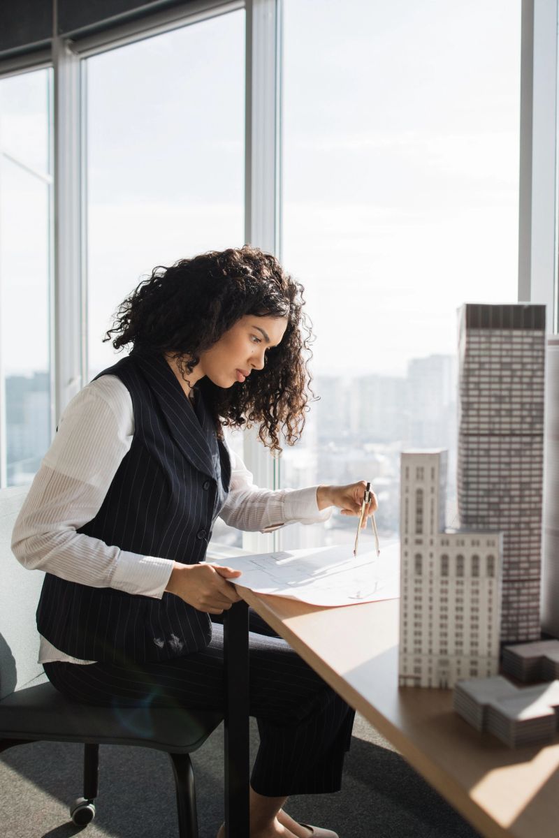 A designer studies scale models and technical drawings by a bright window in a high-rise office overlooking the city skyline