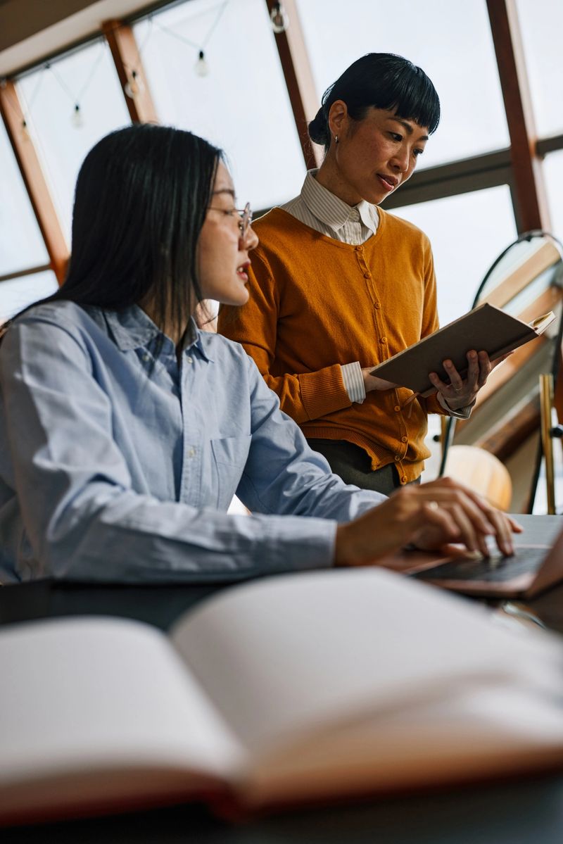 Young Thai woman and middle-aged Japanese colleague discuss a business project at a modern office desk, using a laptop and notebook to plan, learn and collaborate with focused, supportive teamwork.