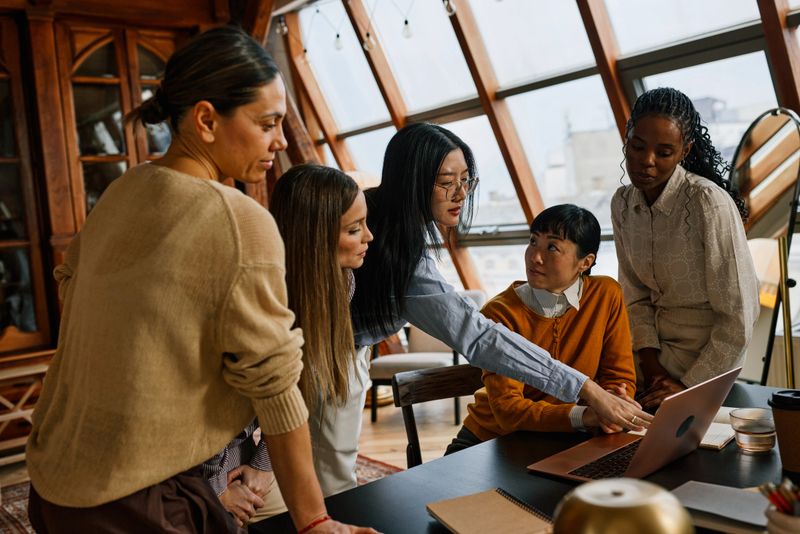 Multiracial women collaborate around a laptop in a warm, sunlit office, discussing a project strategy and sharing ideas. The atmosphere is focused, supportive and creative during an informal business meeting.