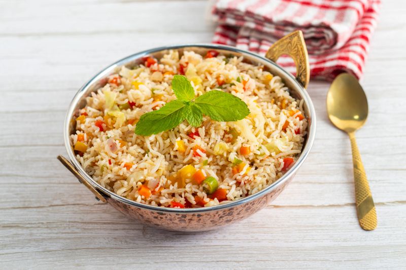 vegetable fried rice in decorative bowl, garnished with mint, served with spoon on wooden background.