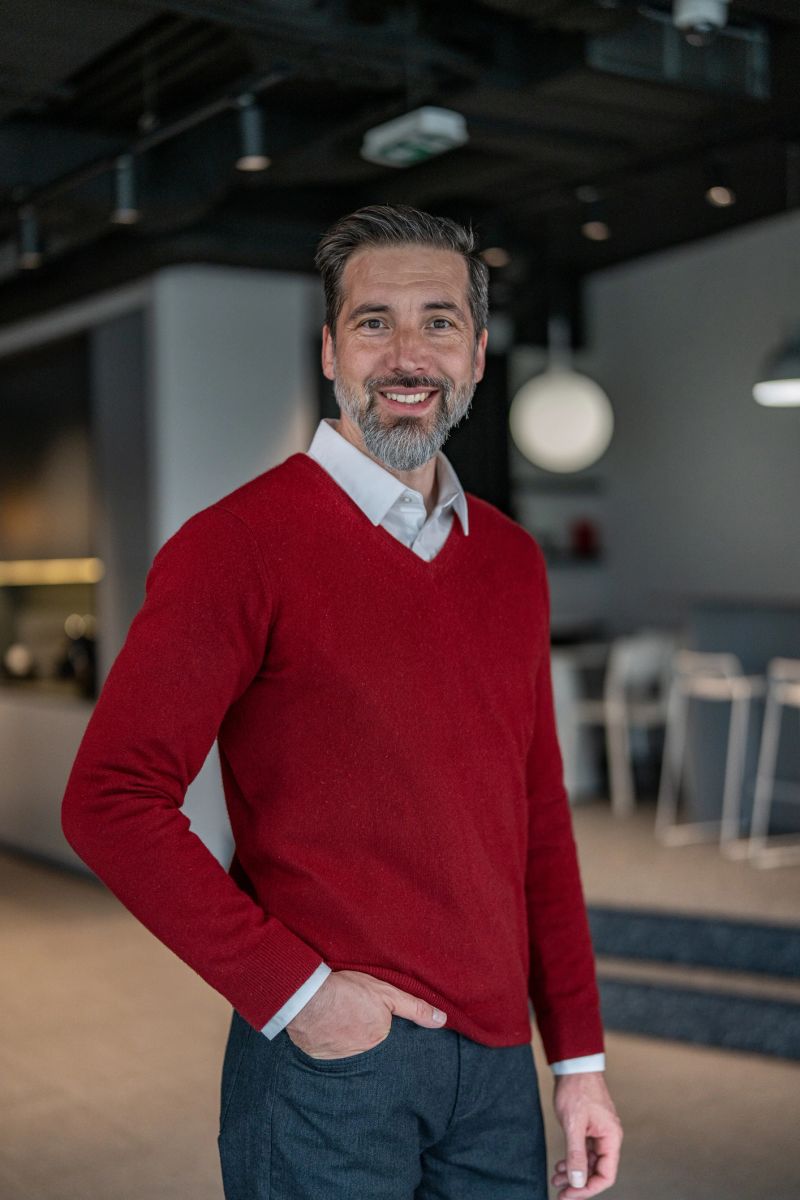 Smiling bearded man in his 40s wearing a red sweater and white collar, standing confidently with hands in pockets in a modern coworking office, looking at camera, relaxed and professional