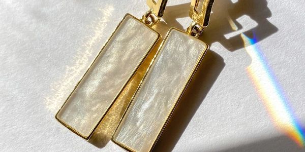 Gold and pearl rectangular drop earrings casting shadows and a rainbow on white surface.