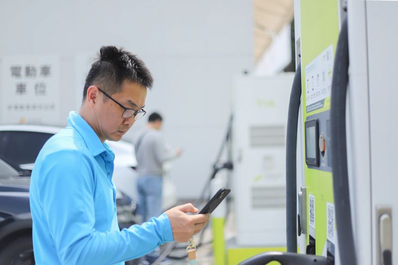 A middle-aged Asian man charges his electric vehicle at a charging station, paying electronically with his smartphone. Electric vehicles have become increasingly popular in recent years and are also supported by environmentalists. The discussion revolves around electronic payment, transportation, and environmental protection issues, and sustainable lifestyles.