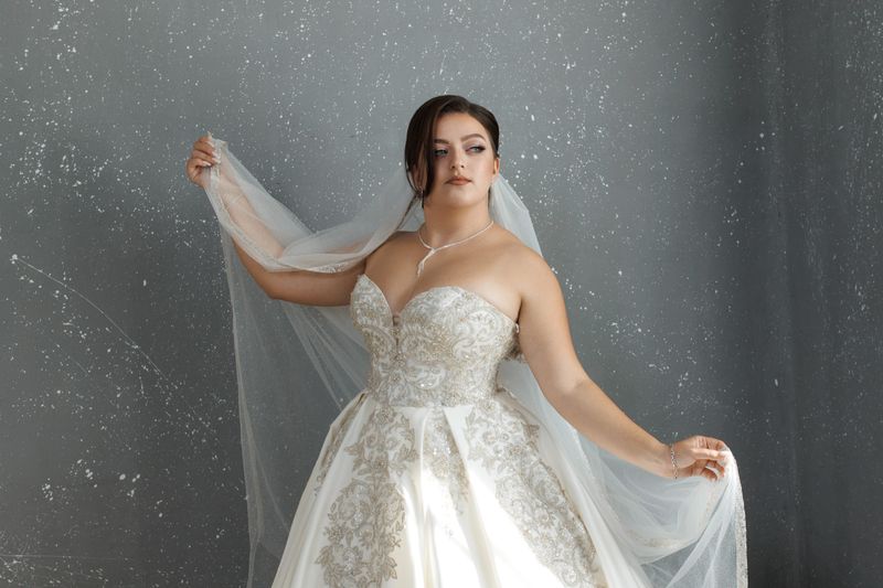 Bride wears a stunning gown with elegant lace details, holding her veil gracefully in a modern indoor space. The soft lighting highlights her joyful expression and the beauty of her attire.
