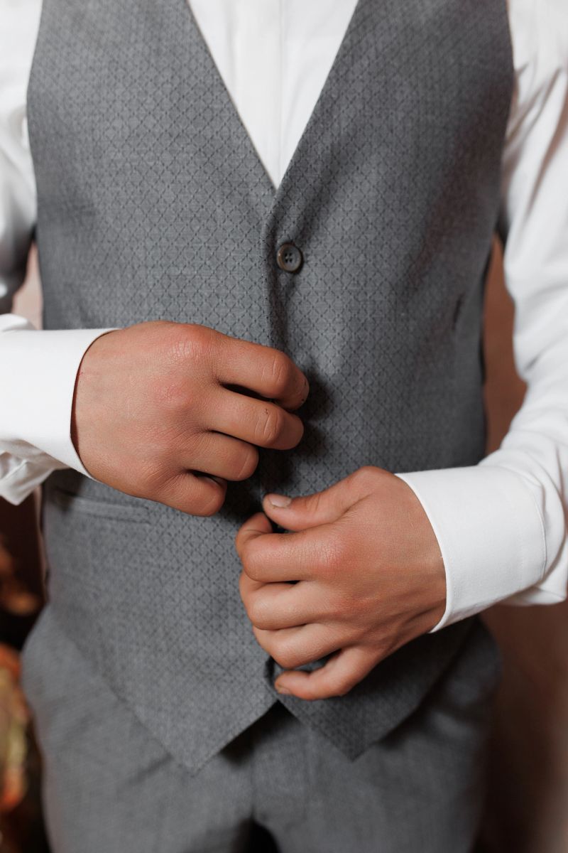 A groom in a stylish gray vest is adjusting his outfit while preparing for his wedding ceremony, showcasing a moment of anticipation and excitement in a beautifully decorated venue.