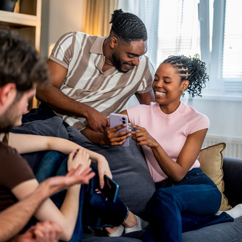 Young diverse students sitting on a sofa, smiling and looking at a smartphone screen, enjoying digital content together