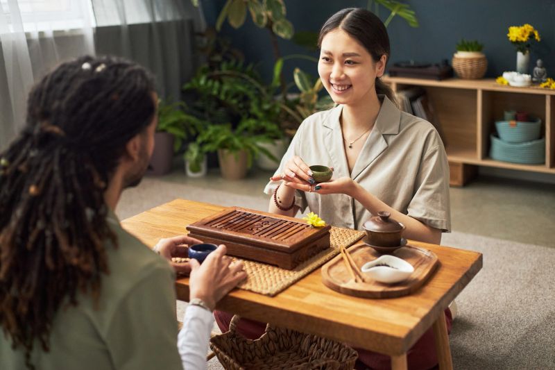 Young Asian woman and young Black man participating in traditional tea ceremony, sitting at wooden table with tea set, smiling and engaging in cultural activity indoors