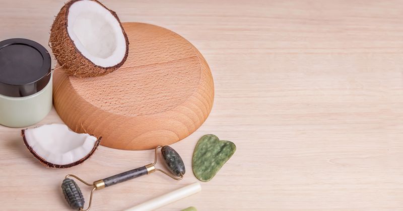 Coconut halves, a jade roller, and skincare products are arranged on a wooden surface, showcasing natural beauty and wellness items in a clean setting