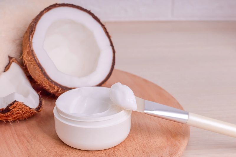 A half coconut shell beside a jar of cosmetic cream and a brush on a wooden surface, showcasing natural beauty products and ingredients