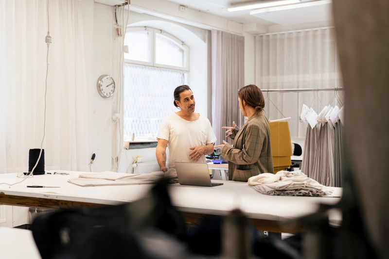 Designer and tailor confer in a bright textile workshop, reviewing fabric samples, patterns and production plans while using a laptop and garments on a rack to coordinate a clothing collection.