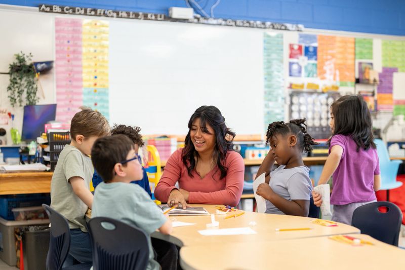 A smiling elementary teacher leads a small group of diverse students around a table, engaging them in hands-on learning and collaborative activities in a colorful, well-organized classroom environment.