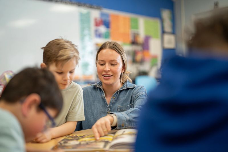 Elementary teacher guides young students through a picture book during a collaborative reading activity in a colorful classroom, fostering literacy, engagement, and supportive student-teacher interaction.