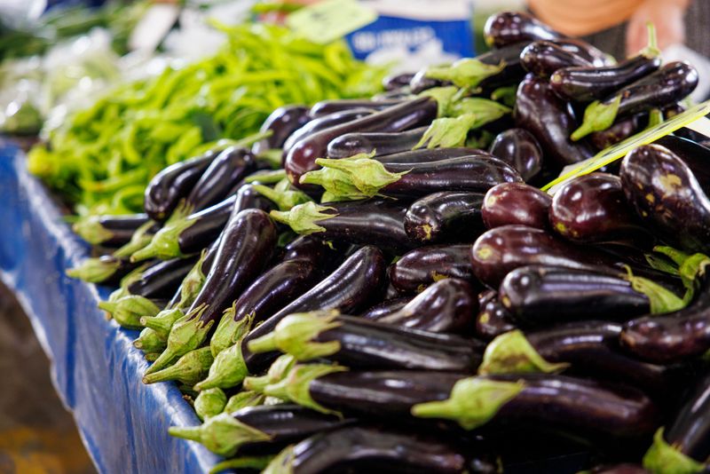 Fresh eggplants displayed at a local open-air market stall in Türkiye, with long green peppers visible in the background. Shiny purple aubergines with green stems are arranged in abundance, showcasing seasonal vegetables commonly used in Mediterranean and Turkish cuisine. The image reflects everyday shopping at a traditional farmers market, highlighting freshness, healthy food choices, and locally grown produce in a vibrant market environment.
